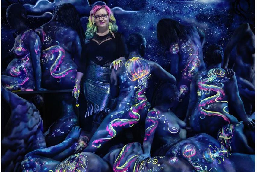 Woman standing with phosphorescent paint brush in hand in a dark lit room, with naked bodies posing around her, painted with jelly fish and flowing patterns.