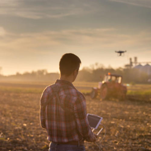Man standing in field controlling a drone by remote control, hovering over a ploughing tractor