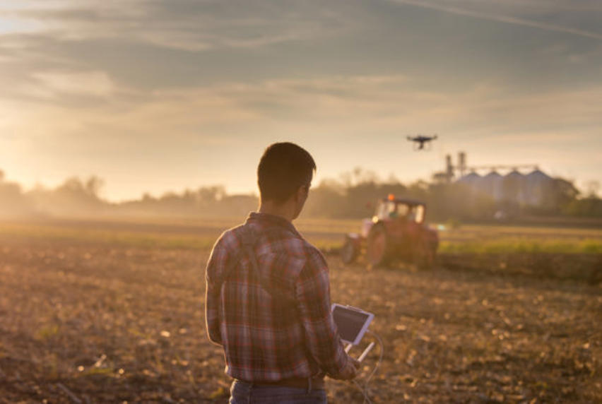 Man standing in field controlling a drone by remote control, hovering over a ploughing tractor