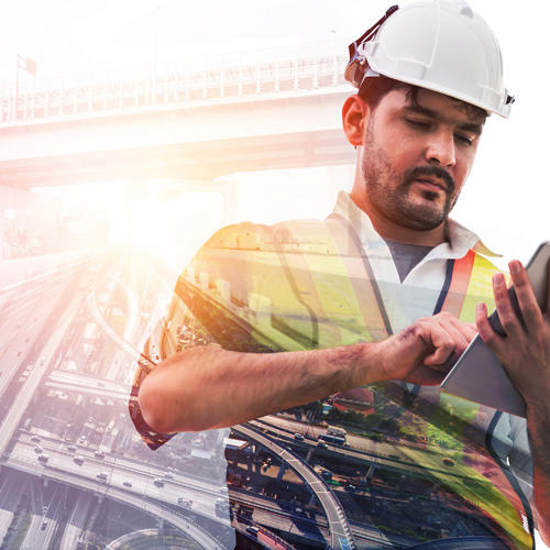 Man with construction hat, hi-vis vest, working on a tablet device, with an image of freeways and a bridge in background