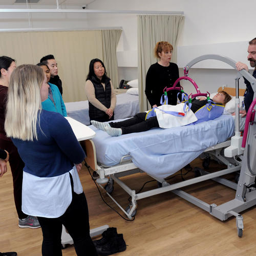 An image of a group of Aged Care students gathered around a patient lying in a hospital bed with a teacher showing how to use a patient hoist.