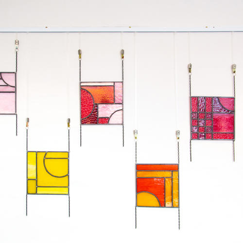An image of five small coloured stained glass panels hanging on chains against a white background.